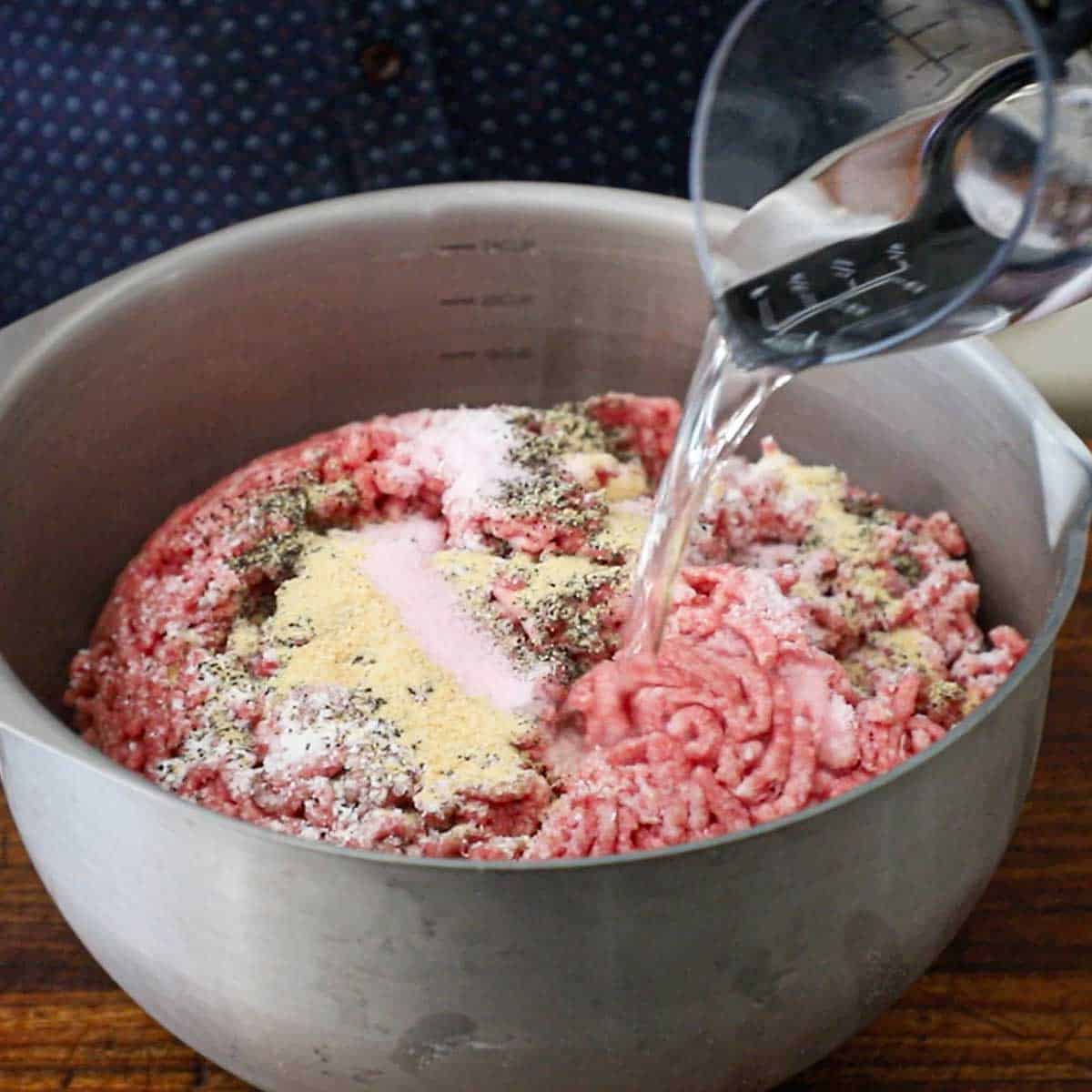 A person pouring water from a plastic measuring up into a large metal bowl filled with freshly ground beef and pork and topped with seasonings and pink curing salt.