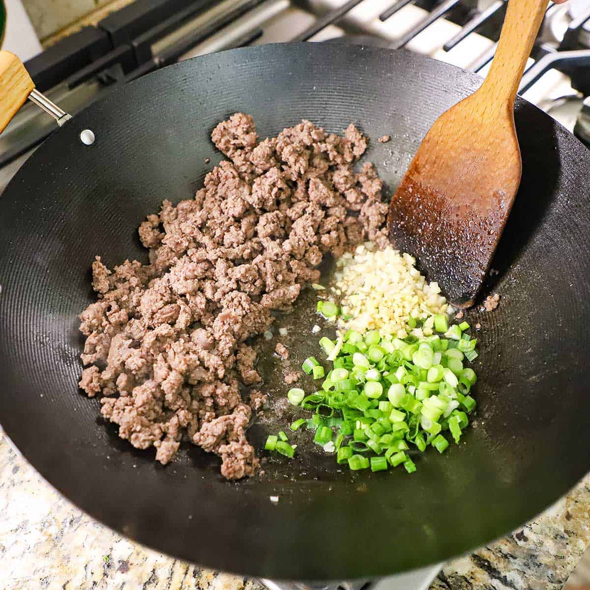 A person using a wooden spatula to stir a mound of minced ginger, garlic, and chopped scallions in oil on one side of a wok with the other side filled with cooked ground beef.