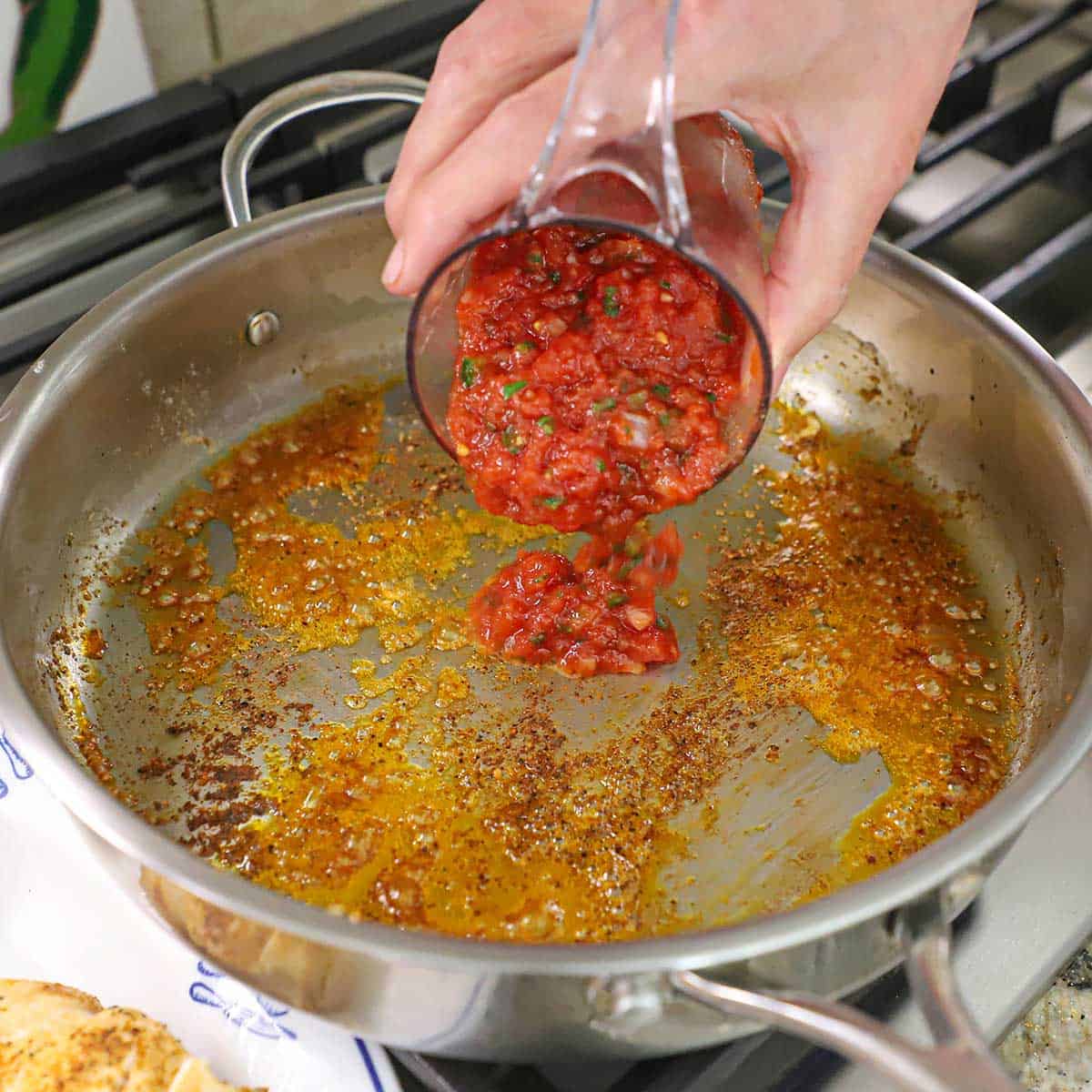 A person pouring homemade salsa into a skillet that has a layer of simmering oil and stuck-on bits from chicken breasts that were previously seared in the skillet.