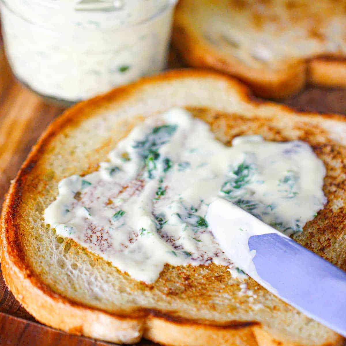 A person using a butter knife to smear garlic basil aioli over one side of a toasted slice of sourdough bread.