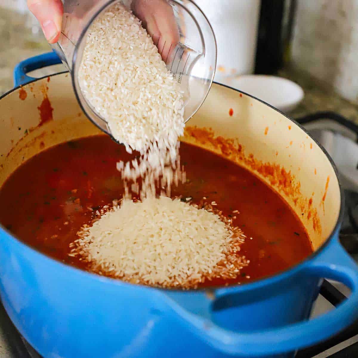 A person pouring uncooked long-grain rice from a plastic measuring cup into a large Dutch oven that is filled with a seasoned tomato sauce.