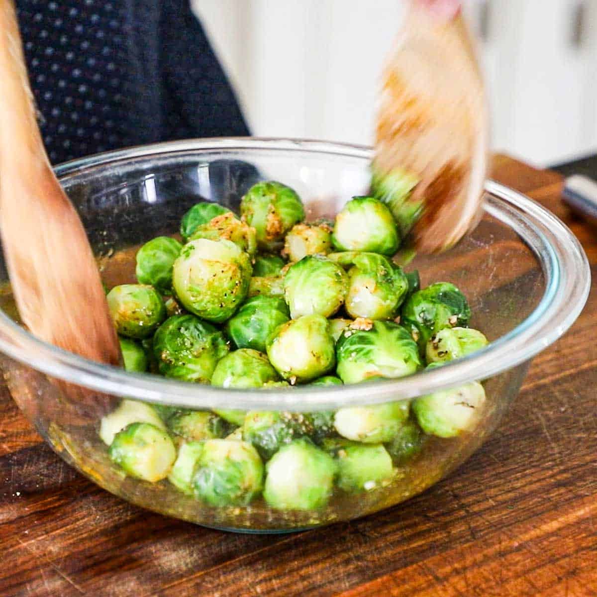 A person using two wooden spoons to toss par-boiled Brussels sprouts in a large glass bowl with melted butter, olive oil, and seasonings.
