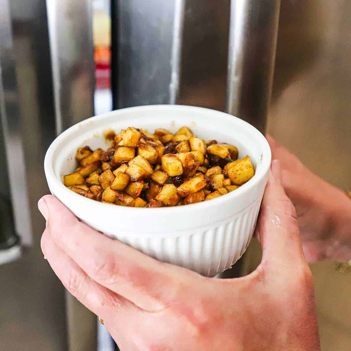 A person holding a large white ramekin in one hand filled with an apple filling and using his other hand to open the door of a refrigerator.