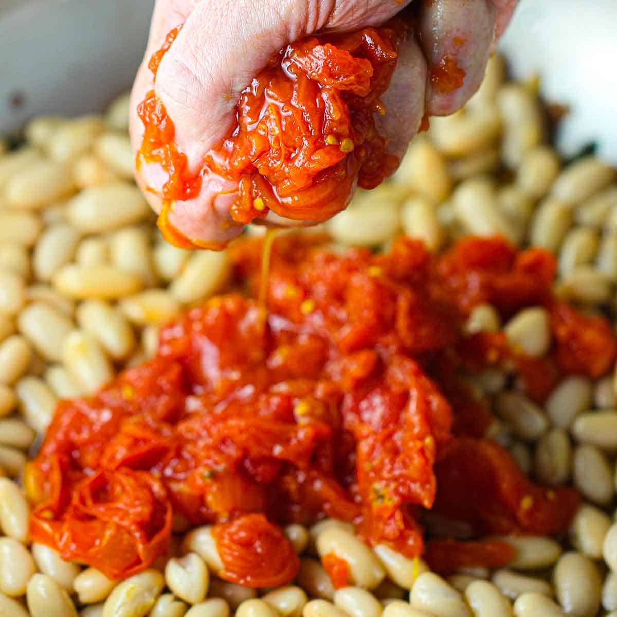 A person crushing canned whole tomatoes over white beans that are being simmer in a large skillet.