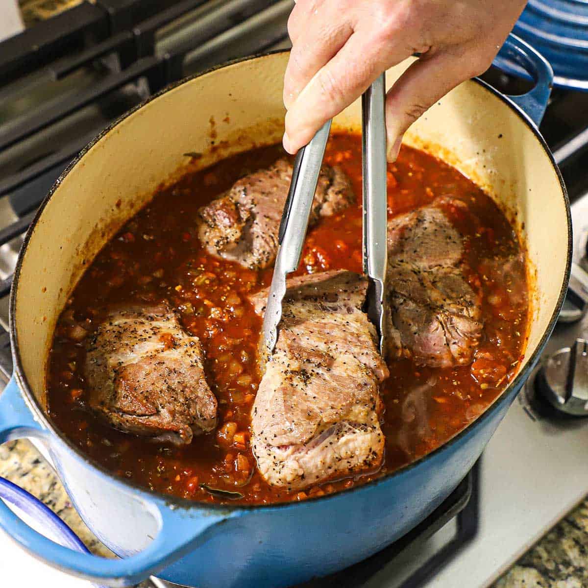 A person nestling chunks of seared pork should into a tomato sauce with vegetables and herbs in a large oval Dutch oven on a gas stove.