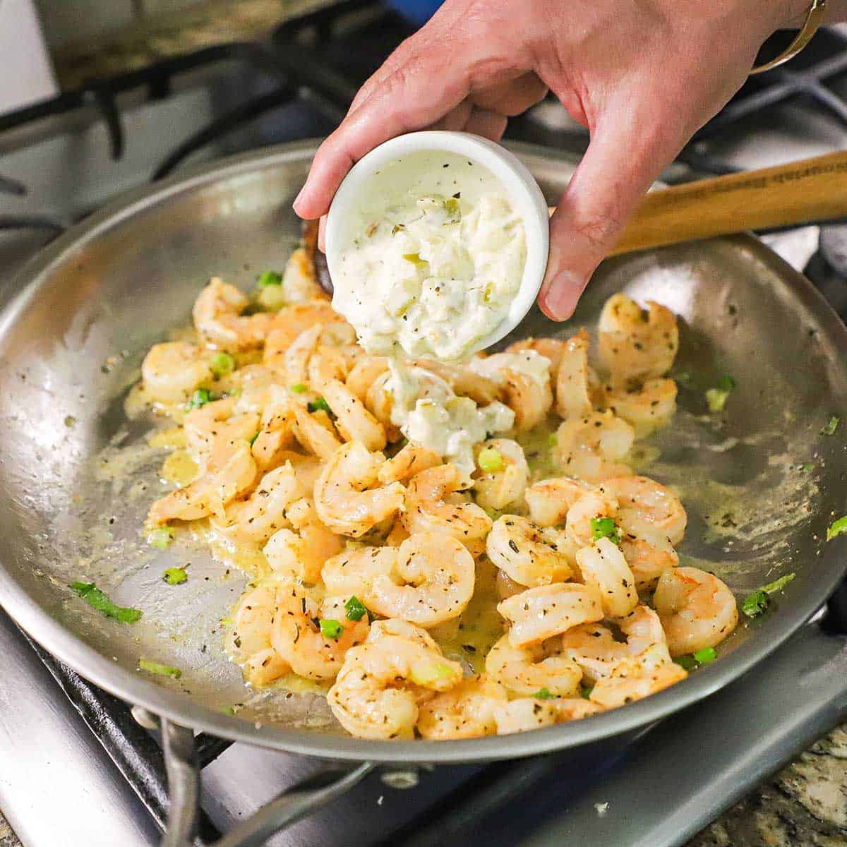 A person pouring a creamy enchilada sauce into a skillet that is filled with pink sautéed shrimp that have been seasoned with salt, pepper, cumin, and chopped scallions.