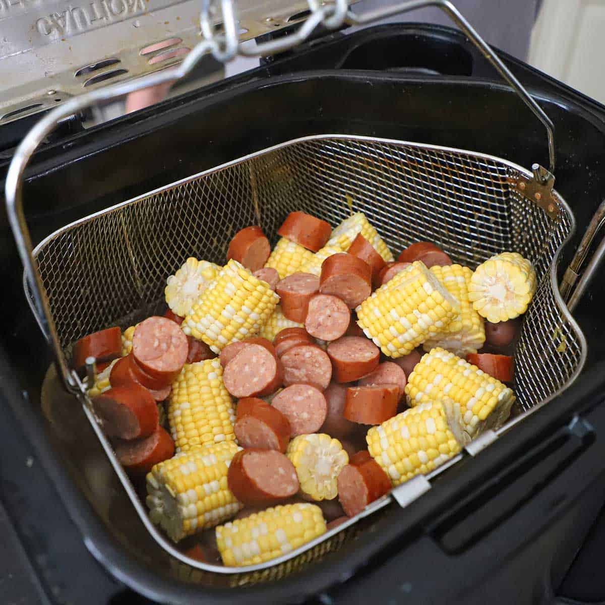 A person lowering a large basket filled with sliced andouille sausage and pieces of fresh corn on the cob into a turkey deep-fryer filled with boiling spiced water.