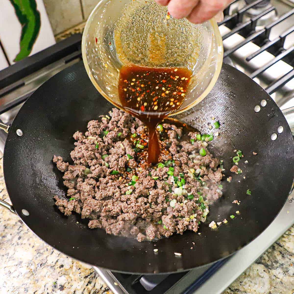 A person pouring a Mongolian beef sauce from a glass bowl into a wok that is filled with simmering ground beef combined with sautéed scallions, ginger, and garlic.