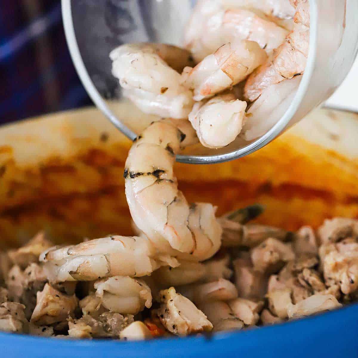 A person transferring uncooked shrimp from a glass bowl into a large Dutch oven the is filled with a simmer Jambalaya sauce.