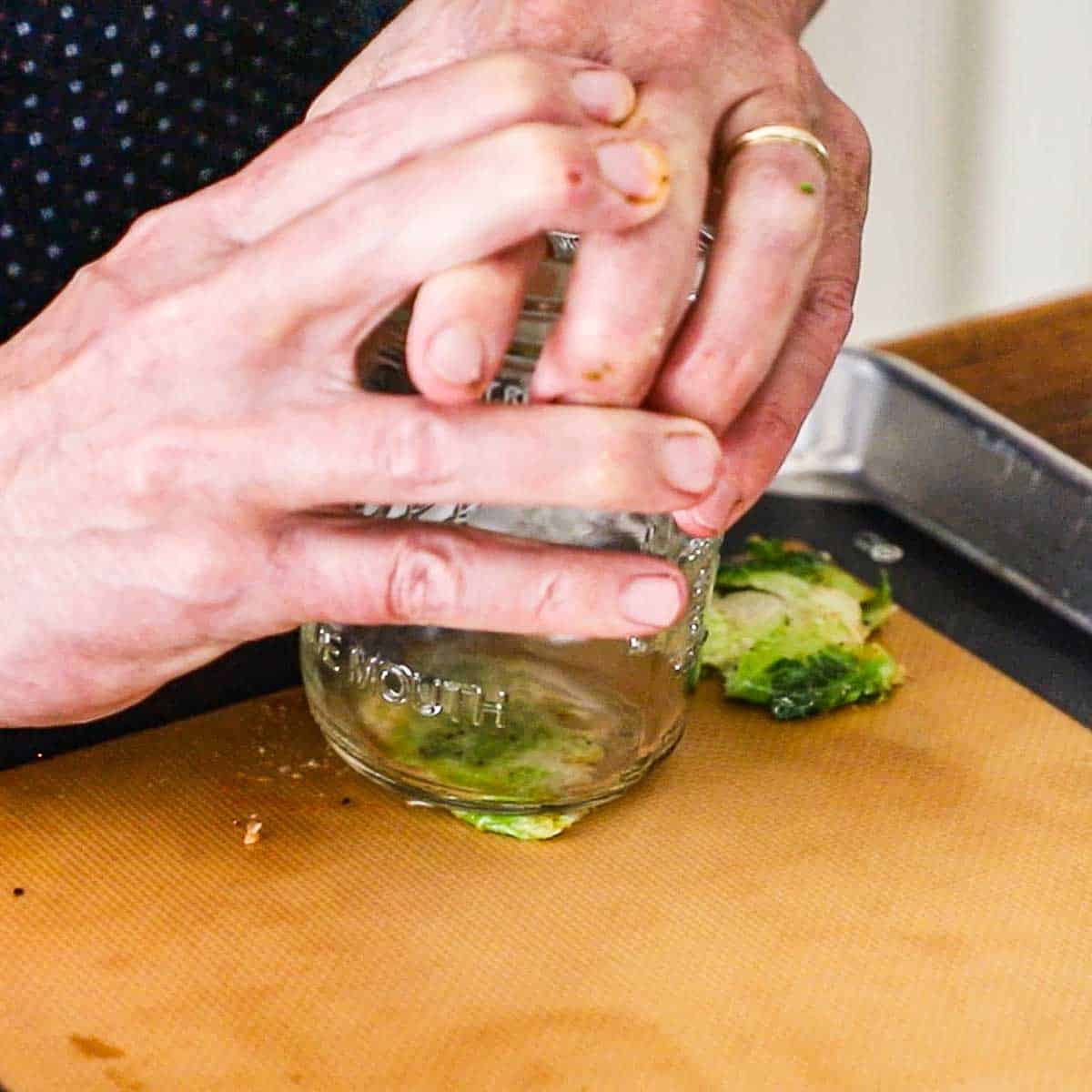 A person using the bottom of a glass jar to flatten a partially cooked Brussels sprout on a sheet pan that is lined with a silicone matt.