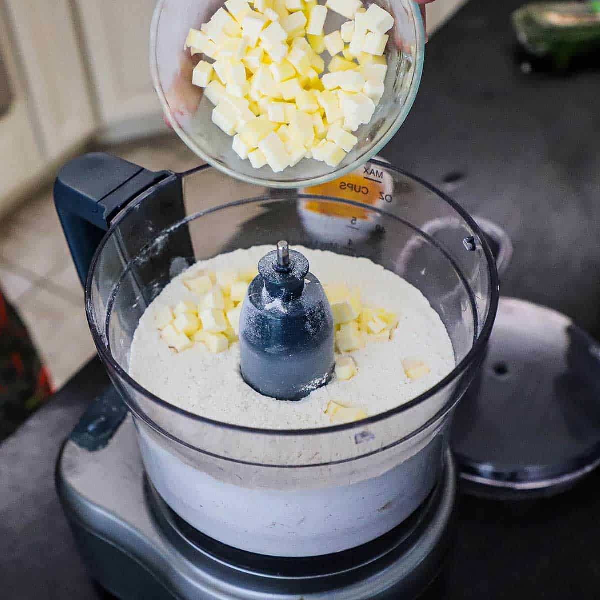 A person dumping chilled cubed butter into a food processor that is filled with all-purpose flour mixed with salt and sugar.