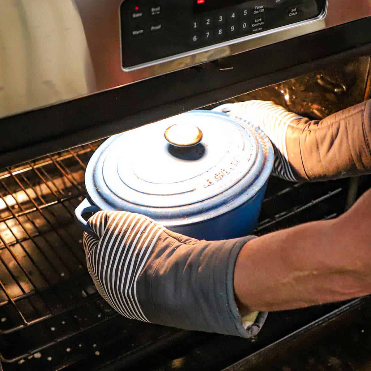A person wearing oven mitts placing a large oval Dutch oven with a lid on it into a preheated oven.