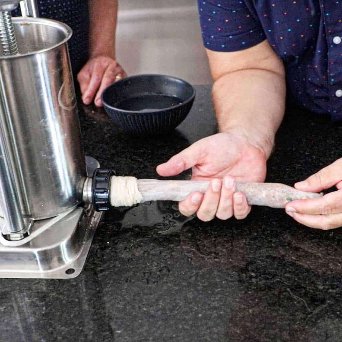 A person using both of his hands to guide a sausage filling being pumped from a sausage pump into natural hog casings on a kitchen counter.