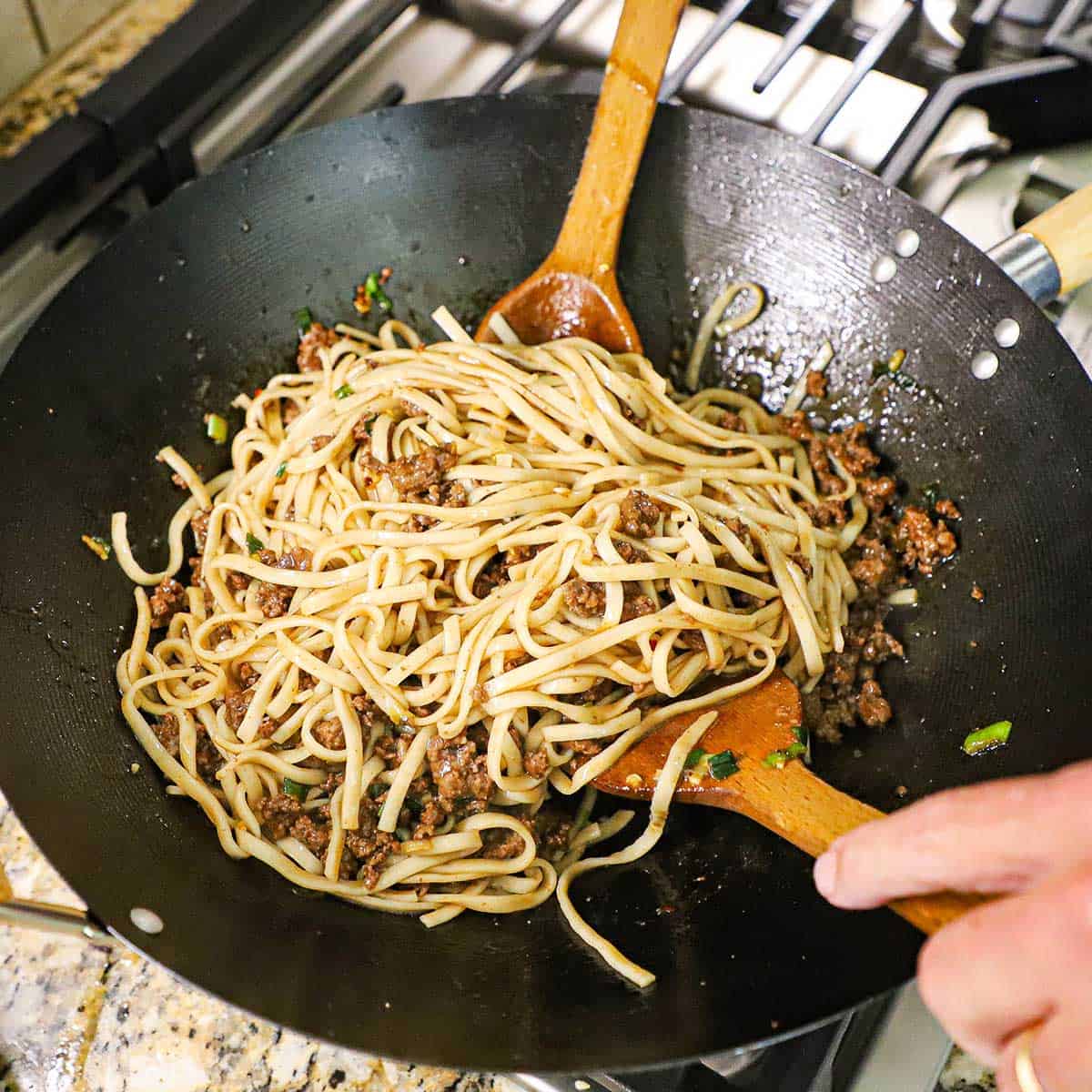 A person using two large wooden spoons to toss cooked udon noodles with Mongolian ground beef in a wok on a gas stove.