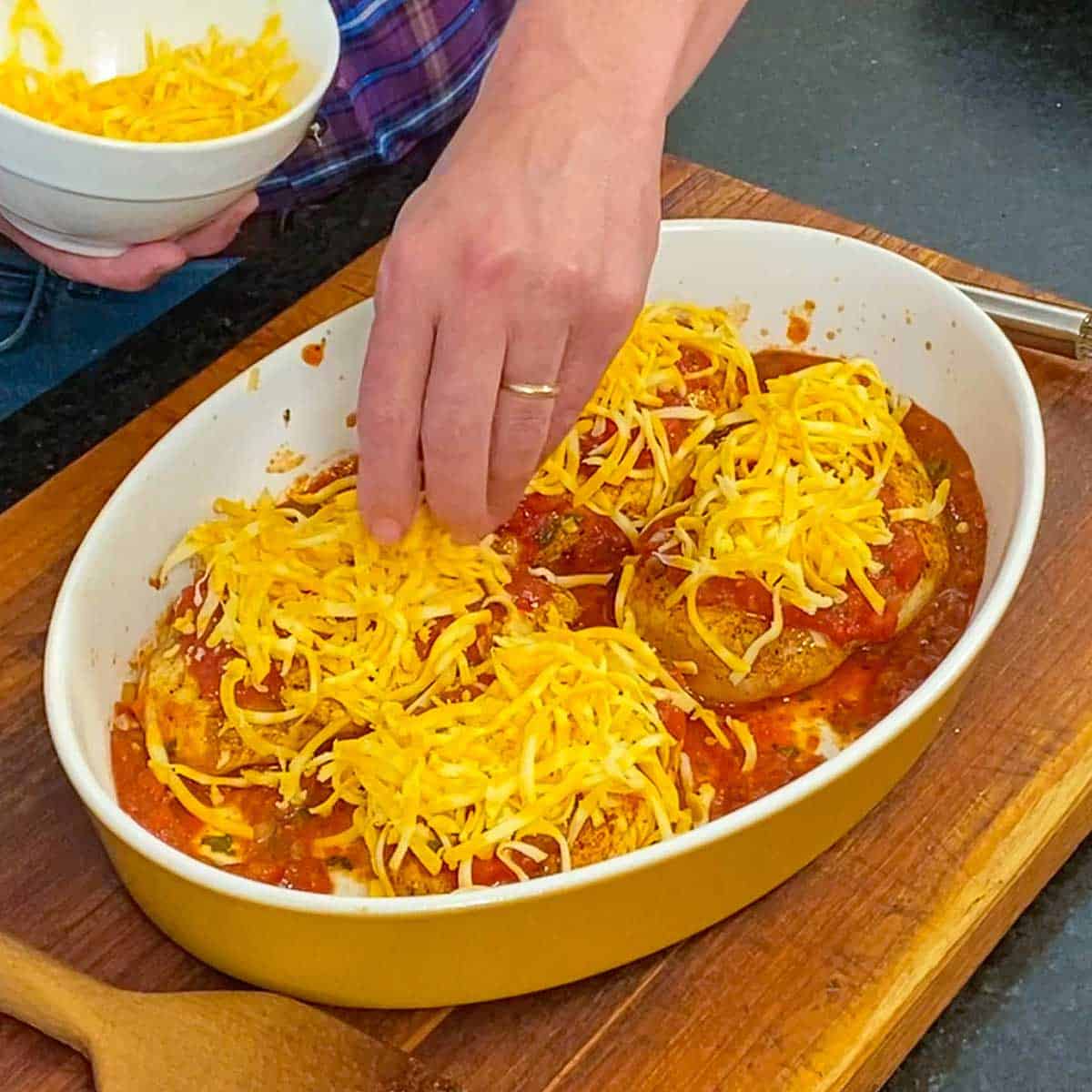 A person placing mounds of shredded Colby Jack cheese over the tops of seasoned seared chicken breasts that also have a layer of salsa over them in a large casserole dish.