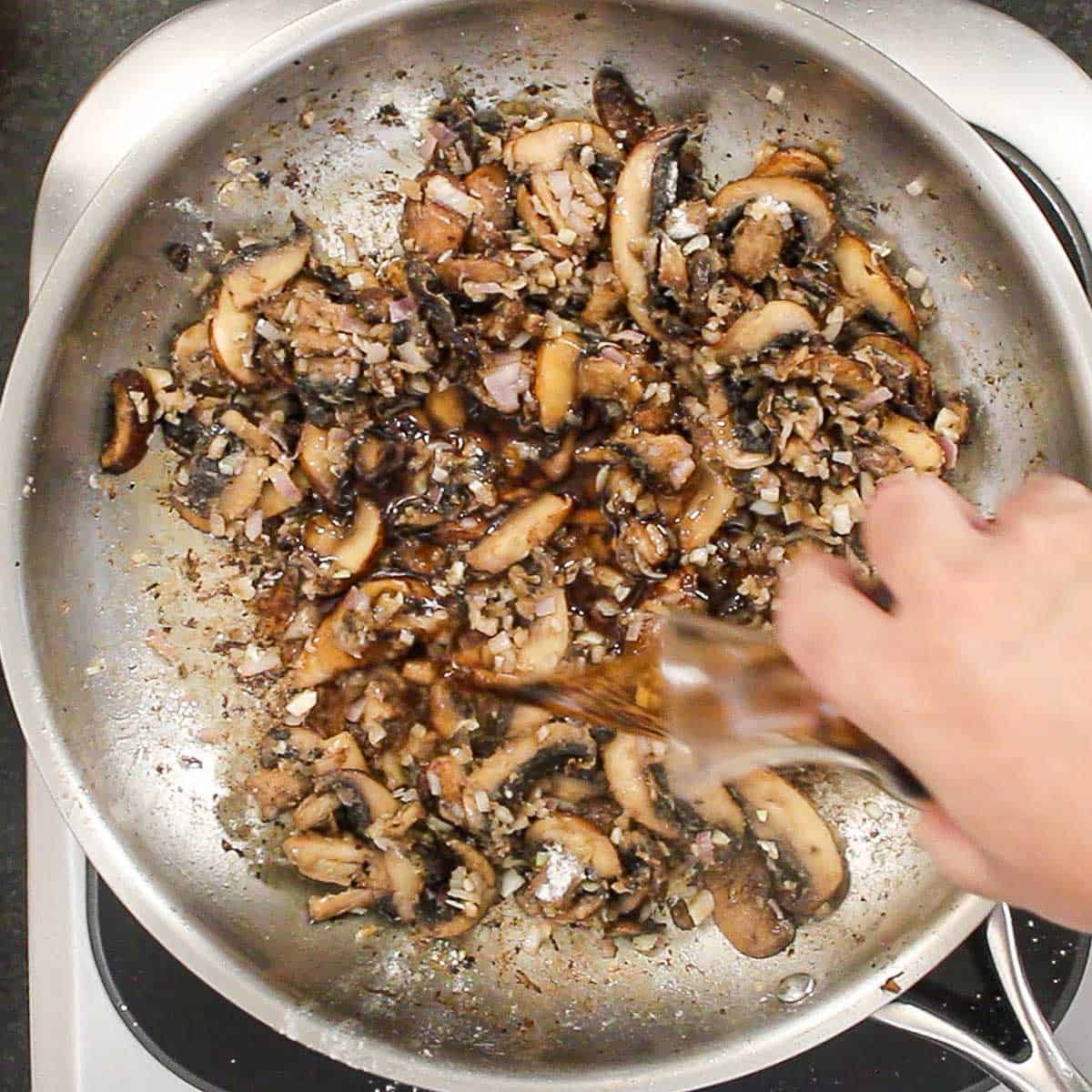 A person pouring Madeira wine from a small glass carafe into a skillet filled with sautéed mushrooms and shallots that have been cooked with flour.