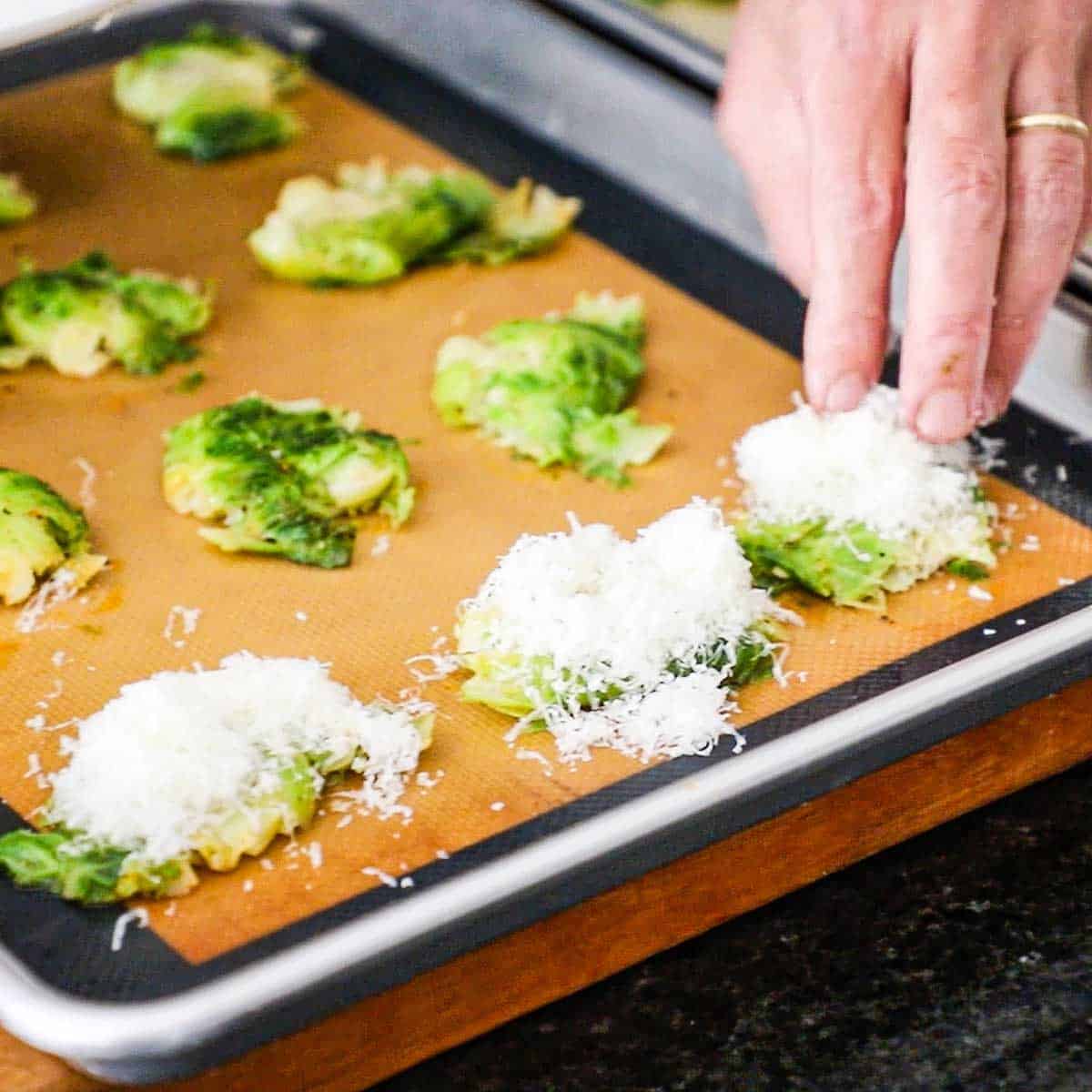 A person using his finger to place a heaping mound of freshly grated Parmesan cheese over the tops of flatten partially Brussels sprouts on a silicone matt in a half sheet pan.