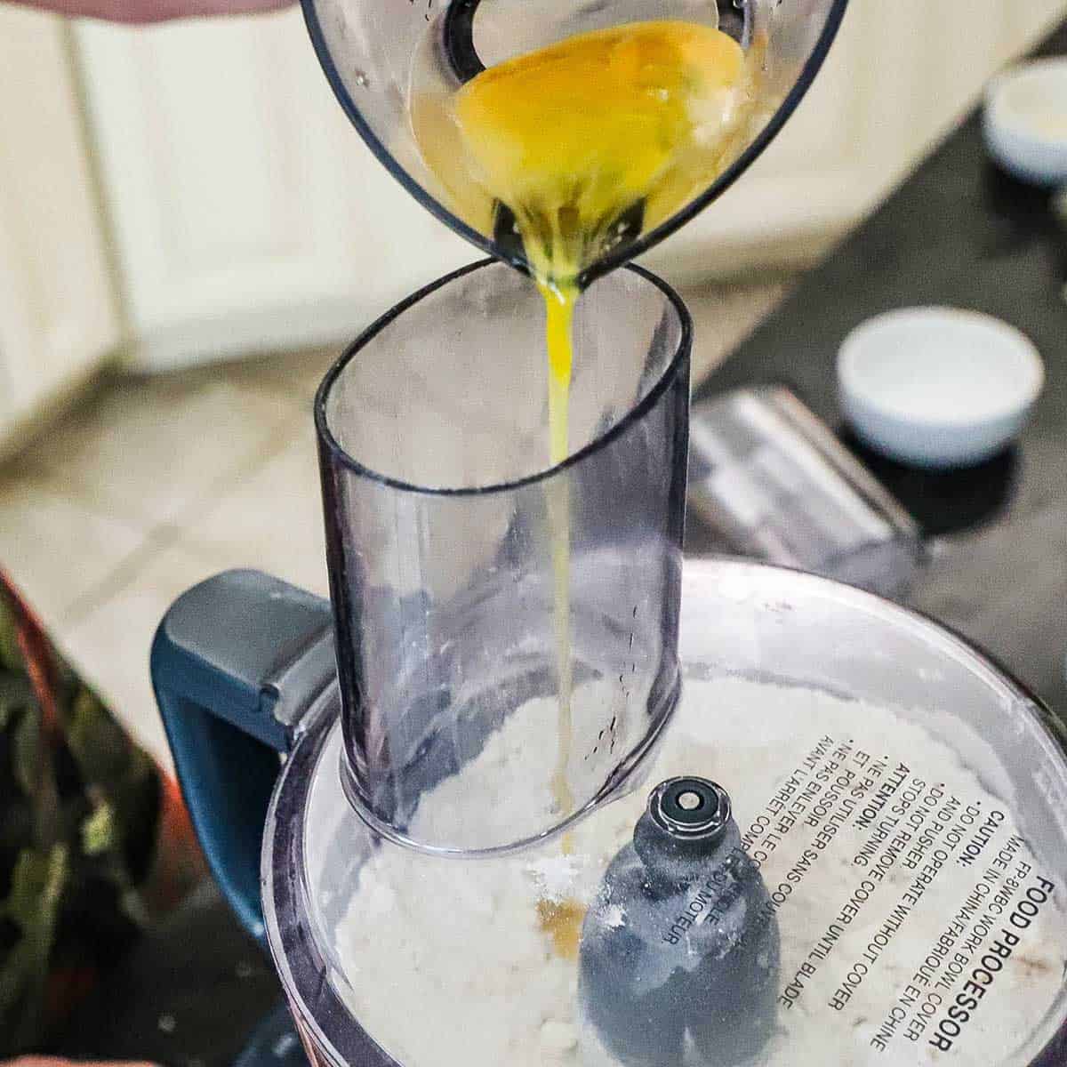 A person pouring an egg and water mixture from a small measuring cup through the tube of a food processor into bowl that is filled with a flour mixture that is being pulsed into dough.