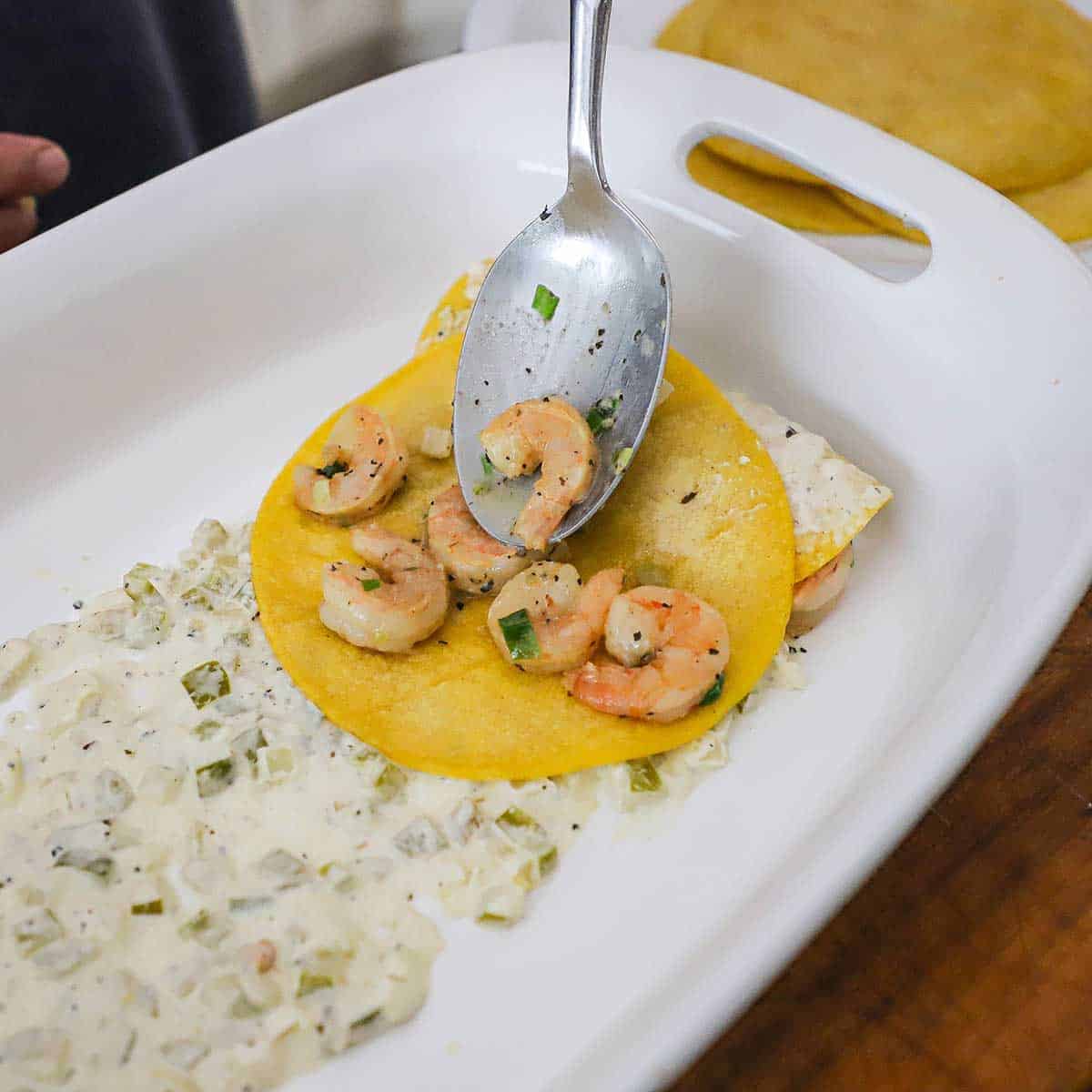 a person using a serving spoon to place cooked seasoned shrimp into the center of a softened corn tortilla that is resting on a creamy enchilada sauce in a baking dish.