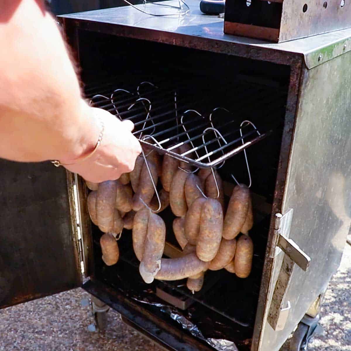A person placing a rack holding rows of hanging uncooked Texas sausage links into the top rung inside an electric smoker.