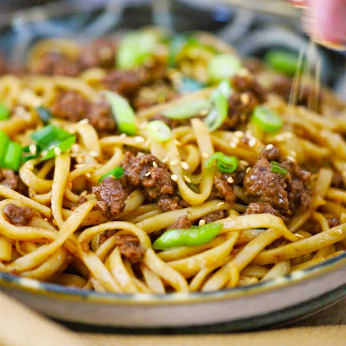 A close-up view Mongolian Ground Beef Noodles that a person is sprinkling toasted sesame seeds over the top of the dish.