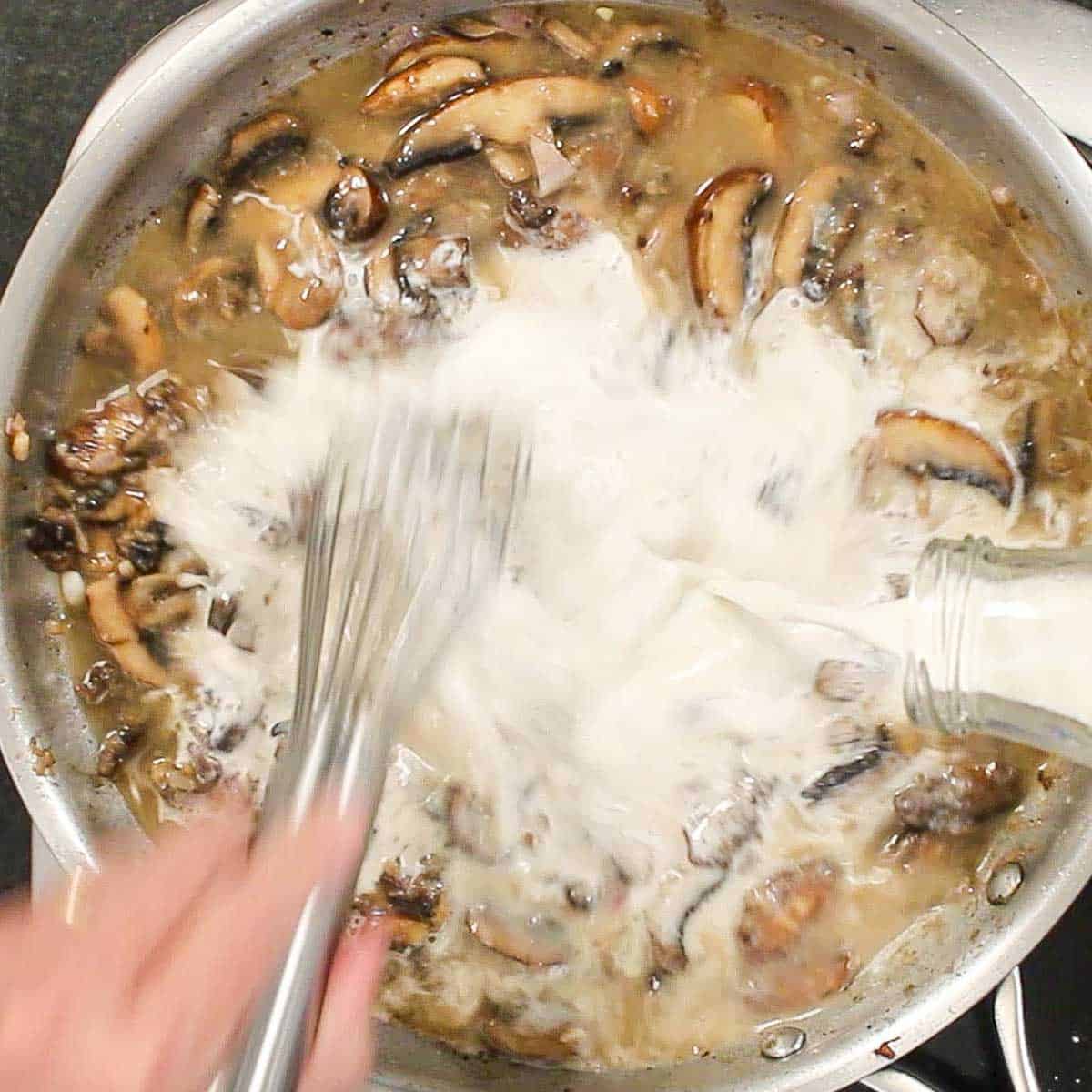 A person pouring heavy cream from a small milk bottle into a skillet that is filled with a creamy brown sauce and sautéed mushrooms.