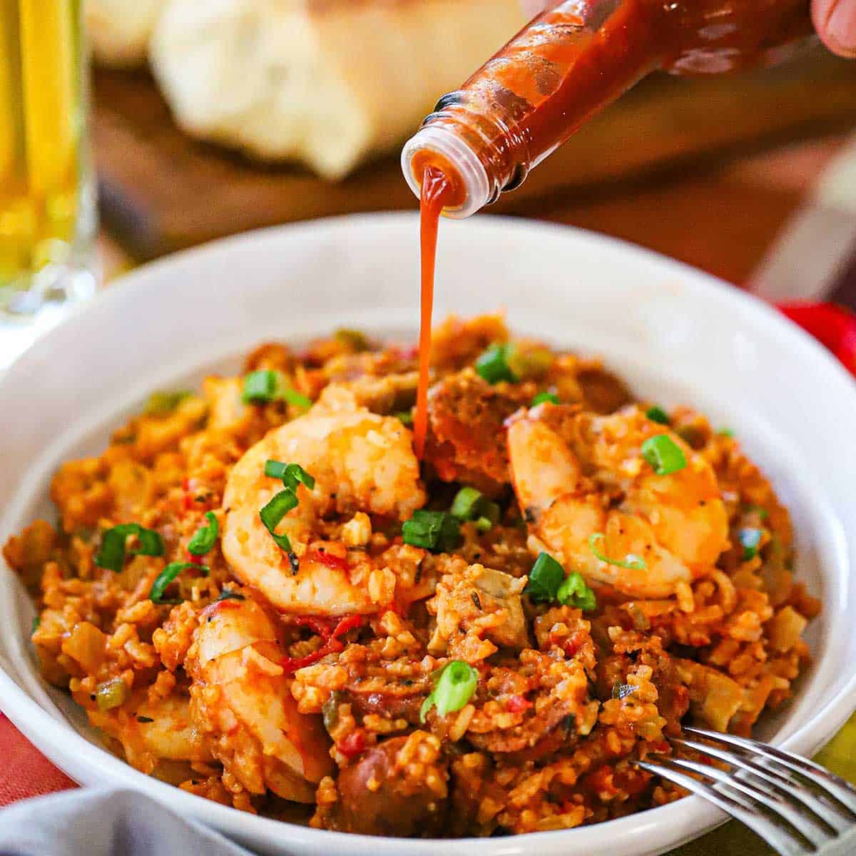 A person pouring Louisiana hot sauce form a bottle onto a serving of authentic jambalaya in a white bowl.