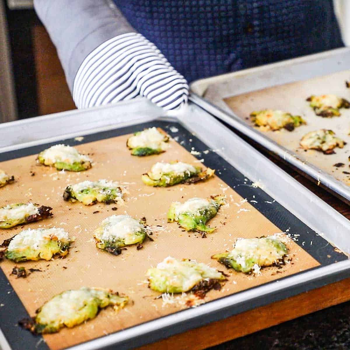 A person wearing oven mitts placing two half sheet pans lined with a silicone matt and topped with freshly baked Parmesan Smashed Brussels Sprouts.