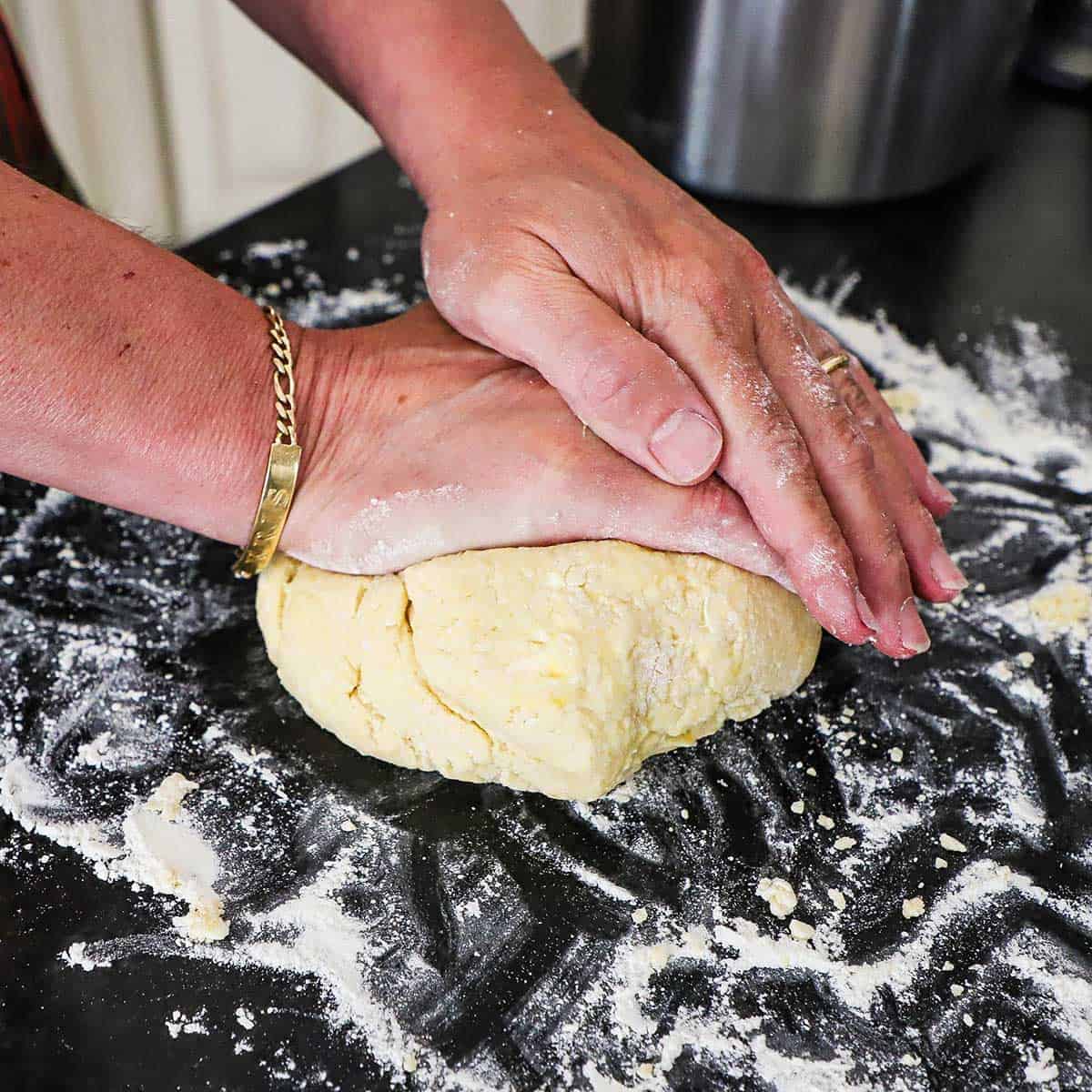 A person using the palms of his hands to knead fruit pie dough on a black marble surface that has been floured all over the surface.