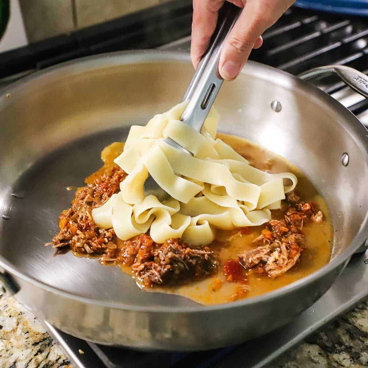 A person using tongs to place cooked pappardelle pasta into a large skillet that contains about 1 cup of braised pork ragu and a splash of pasta water.
