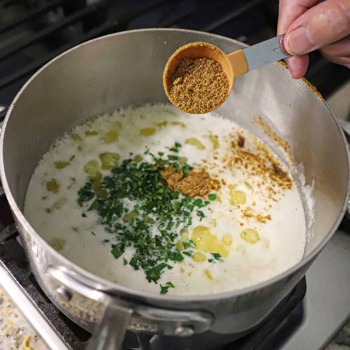 A person adding seafood boil spices from a tablespoon into a saucepan filled with simmering butter, garlic, and chopped Italian parsley.