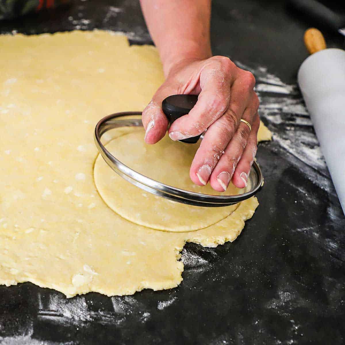 A person using a small lid of a cooking pot to cut a circle of dough from a rolled out diameter of pie dough on a floured surface.