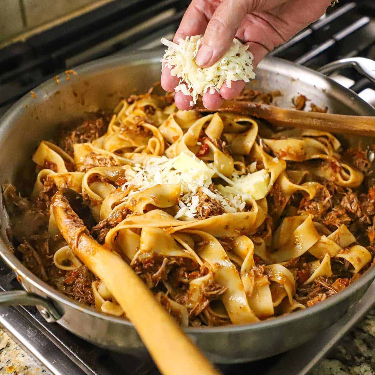 A person using his fingers to place shredded Parmesan cheese into a large skillet filled with slow-braised pork ragu with pappardelle on a gas stove.