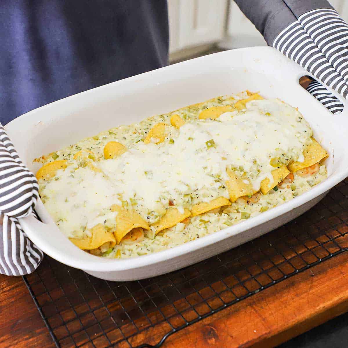 A person wearing oven mitts holding a baking dish that has just been removed from the oven and is filled with bubbling Creamy Shrimp Enchiladas.