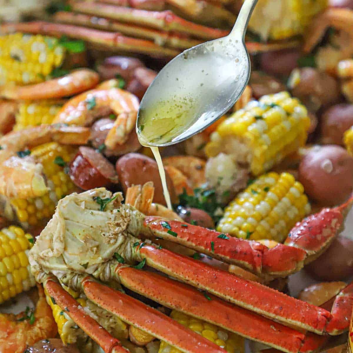 A person using a large spoon to drizzle seasoned melted butter over the top of snow crab legs that are nestled into a large serving of a Louisiana seafood boil.
