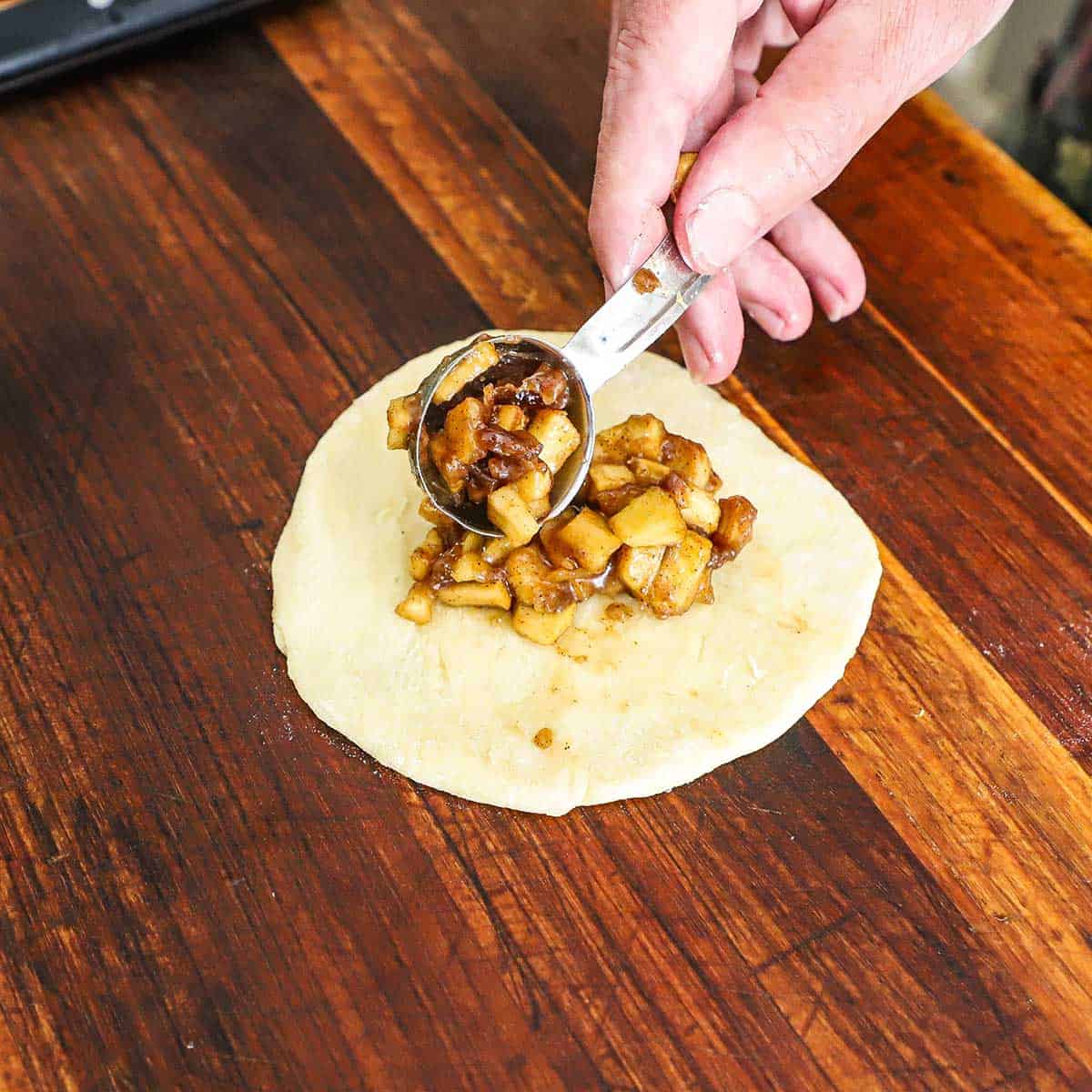 A person using a tablespoon to transfer an apple fruit pie filling onto a circular piece of pie dough on a wooden cutting board.