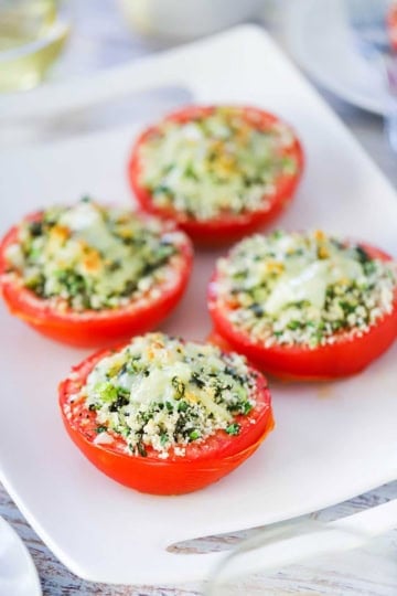 A straight-on view of four baked stuffed summer tomatoes that are topped with lightly browned melted cheese all on a white serving platter.