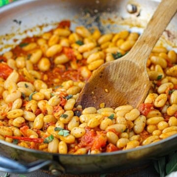 A close-up view of a large silver skillet filled with freshly prepare Sautéed White Beans with Garlic, Sage, and Tomatoes.