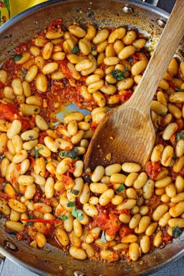 An overhead view of a large silver skillet filled with a freshly prepares Sautéed White Beans with Garlic, Sage, and Tomatoes with a large wooden spoon in the middle.
