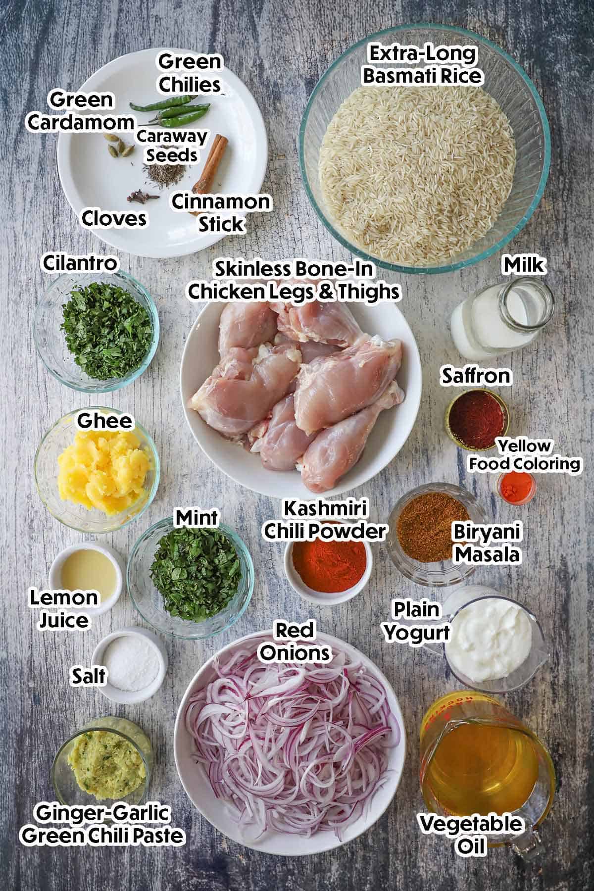 An arrangement of ingredients for Hyderabadi Chicken Biryani on a grey wooden background including raw chicken pieces, basmati rice, red onions, yogurt, ghee, milk, oil, ginger-garlic paste, and spices.