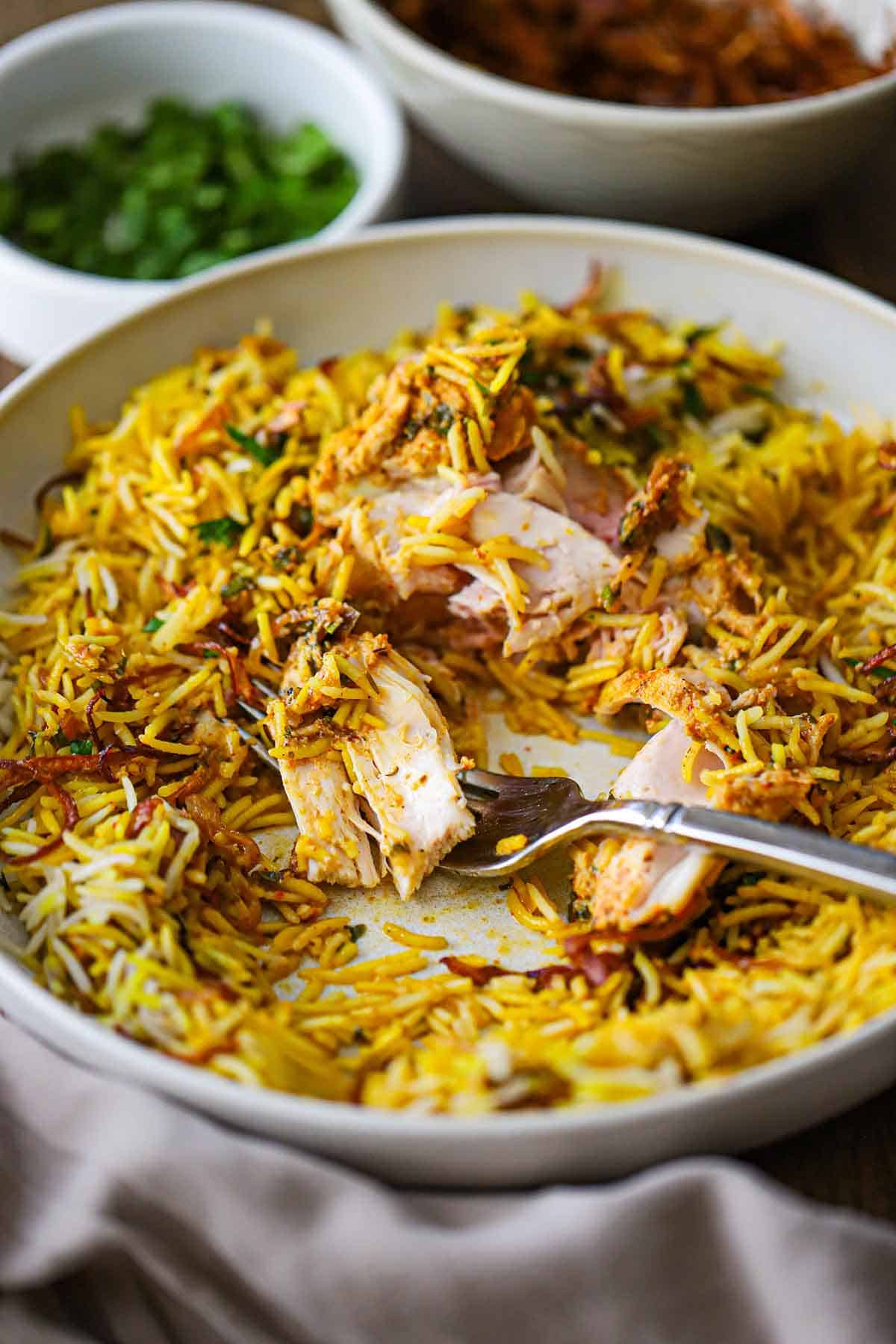 A partially eaten chicken thigh in a shallow bowl filled with a serving of Hyderabadi Chicken Biryani with a fork in the middle of the dish.