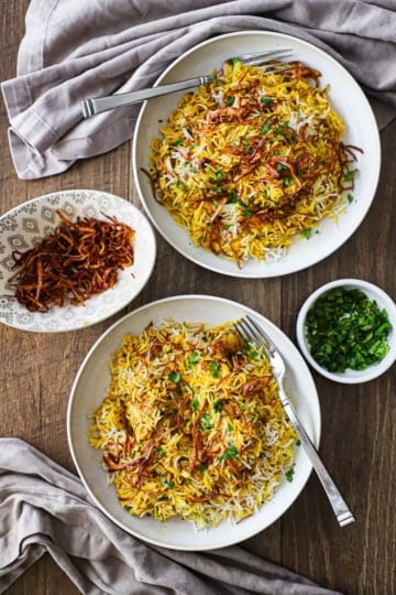 An overhead view of two side-by-side shallow bowls both filled with servings of Hyderabadi Chicken Biryani with a small bowl of fried onions on one side and another small bowl filled with chopped cilantro on the other side.