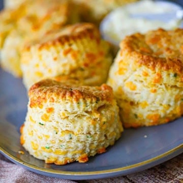 A close-up view of flaky cheddar biscuits sitting in a row on an oval grey platter with a small bowl of softened butter next to the biscuits.