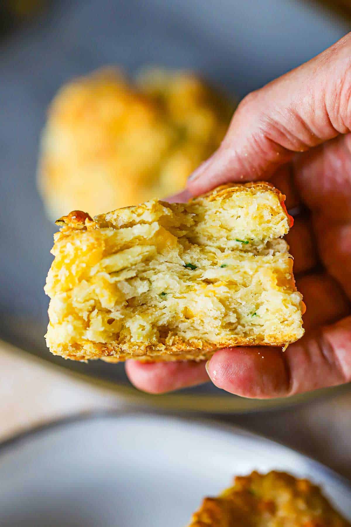A person holding a flaky cheddar biscuit that has been cut in half revealing the layers of bread, cheese, and snipped chives.