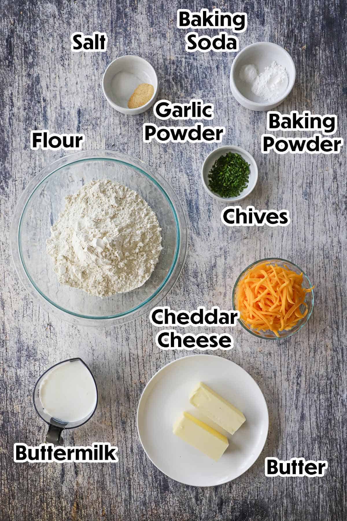 An arrangement of ingredients for flaky cheddar biscuits on a grey wooden background including flour, buttermilk, butter, shredded cheddar cheese, chives, baking powder, baking soda, and seasonings.
