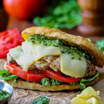 A close-up view of a Grilled Pesto Chicken Sandwich with Smoked Mozzarella on brown paper with potato chips and a bowl of pesto sauce nearby.
