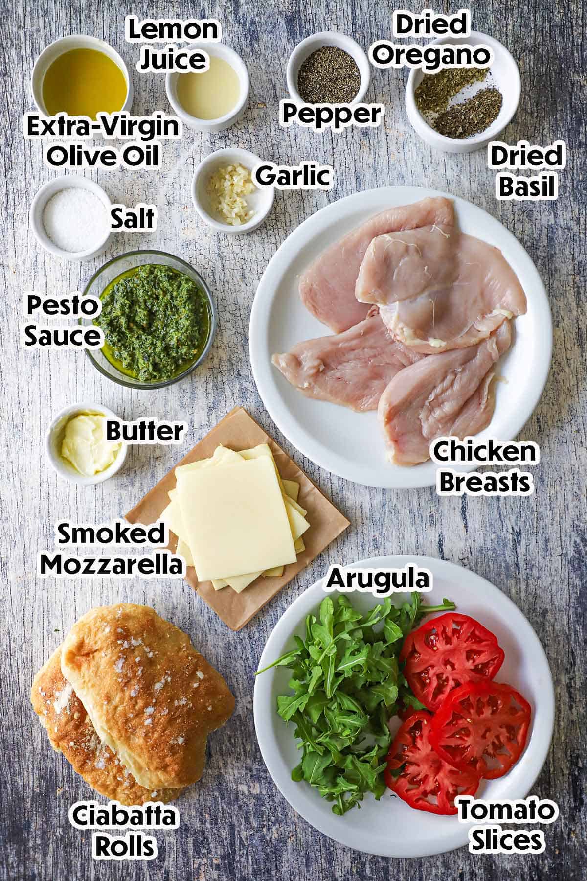 An arrangement of ingredients for Grilled Pesto Chicken Sandwich with Smoked Mozzarella including chicken breast, pesto sauce, cheese slices, ciabatta rolls, arugula, tomato slices, garlic, and seasonings.