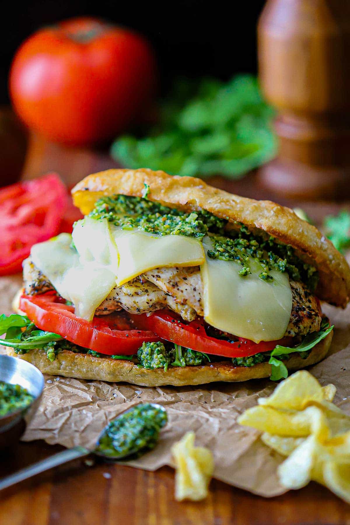 A straight-on view of a Grilled Pesto Chicken Sandwich with Smoked Mozzarella on brown paper with potato chips and a bowl of pesto sauce nearby.