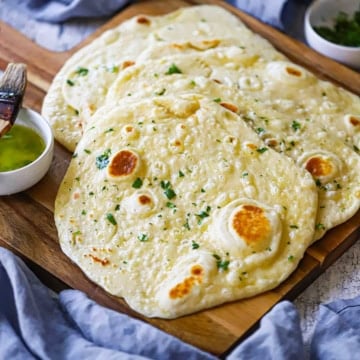 An overhead view of a stack of homemade garlic naan with a small bowl of garlic-cilantro butter nearby as well as a couple of blue cloth napkins.