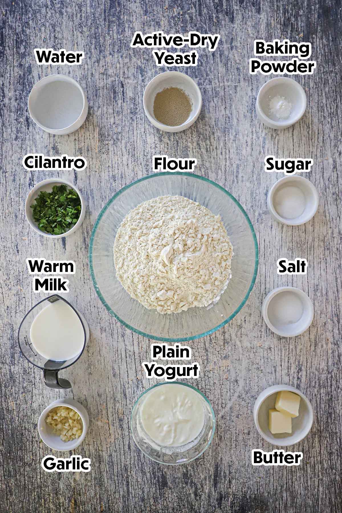 An arrangement of ingredients for Homemade Garlic Naan on a grey wooden background including flour, yeast, baking powder, yogurt, butter, milk, sugar, cilantro, and garlic.